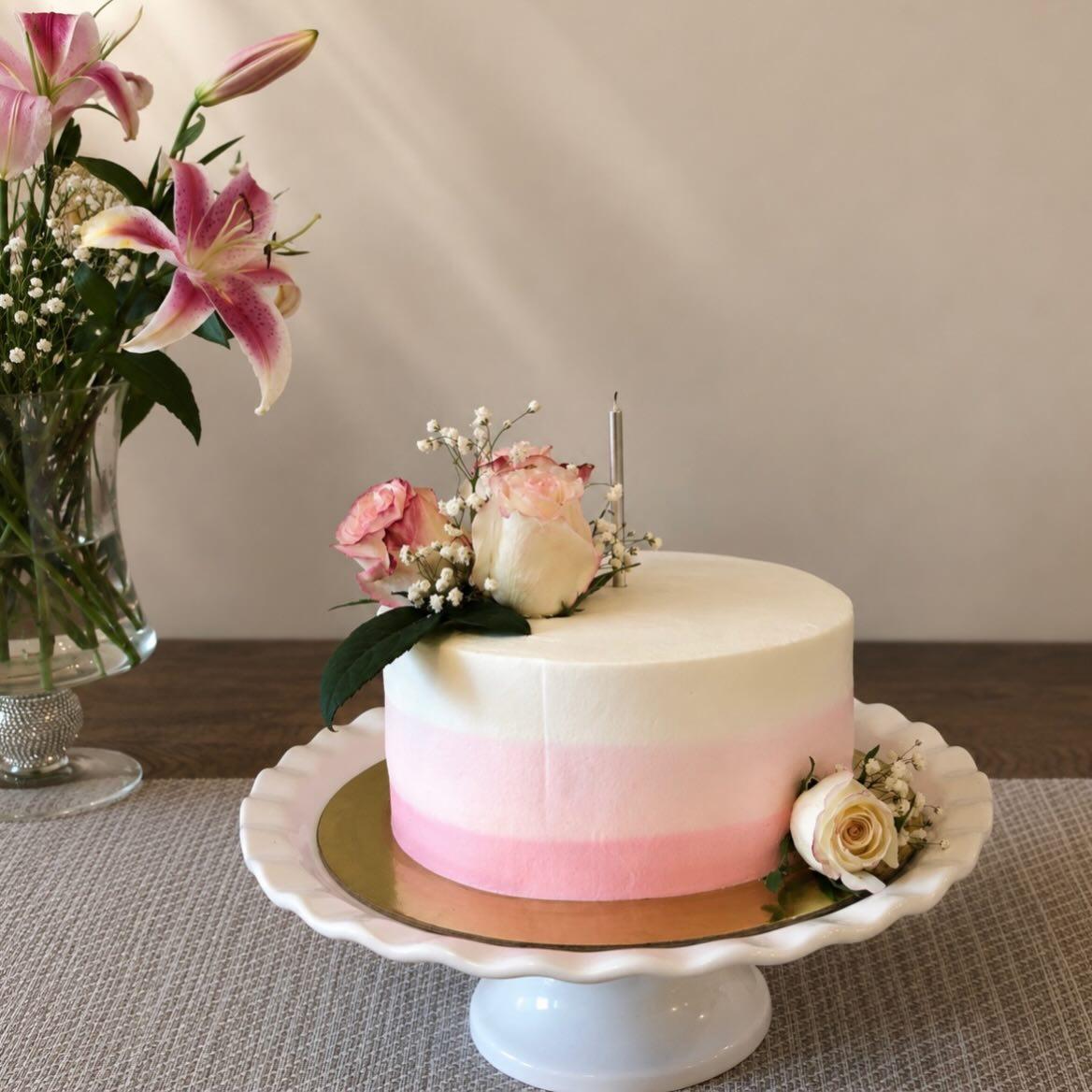 A cake shaded from blush pink to white, topped with garden roses and baby’s breath, with lilies behind