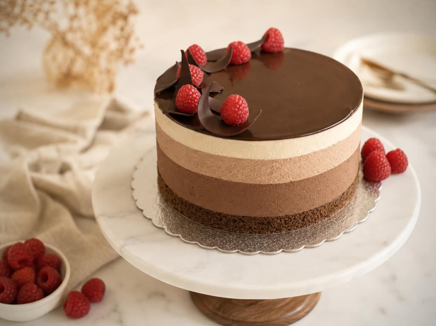 A four-layer chocolate mousse cake with a glossy ganache top, crowned with raspberries and chocolate curls, on a marble stand