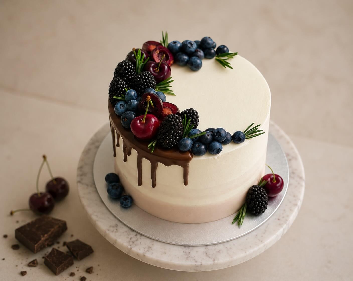 A white cake with a chocolate drip, crowned with a crescent of cherries, blackberries, blueberries and rosemary, on a marble stand