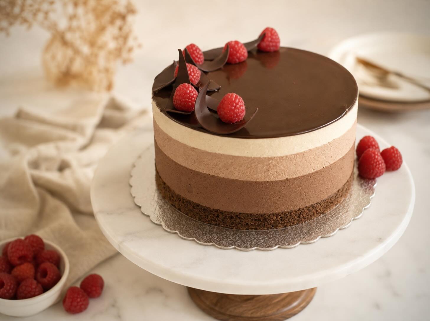 A four-layer chocolate mousse cake with a glossy ganache top, crowned with raspberries and chocolate curls, on a marble stand