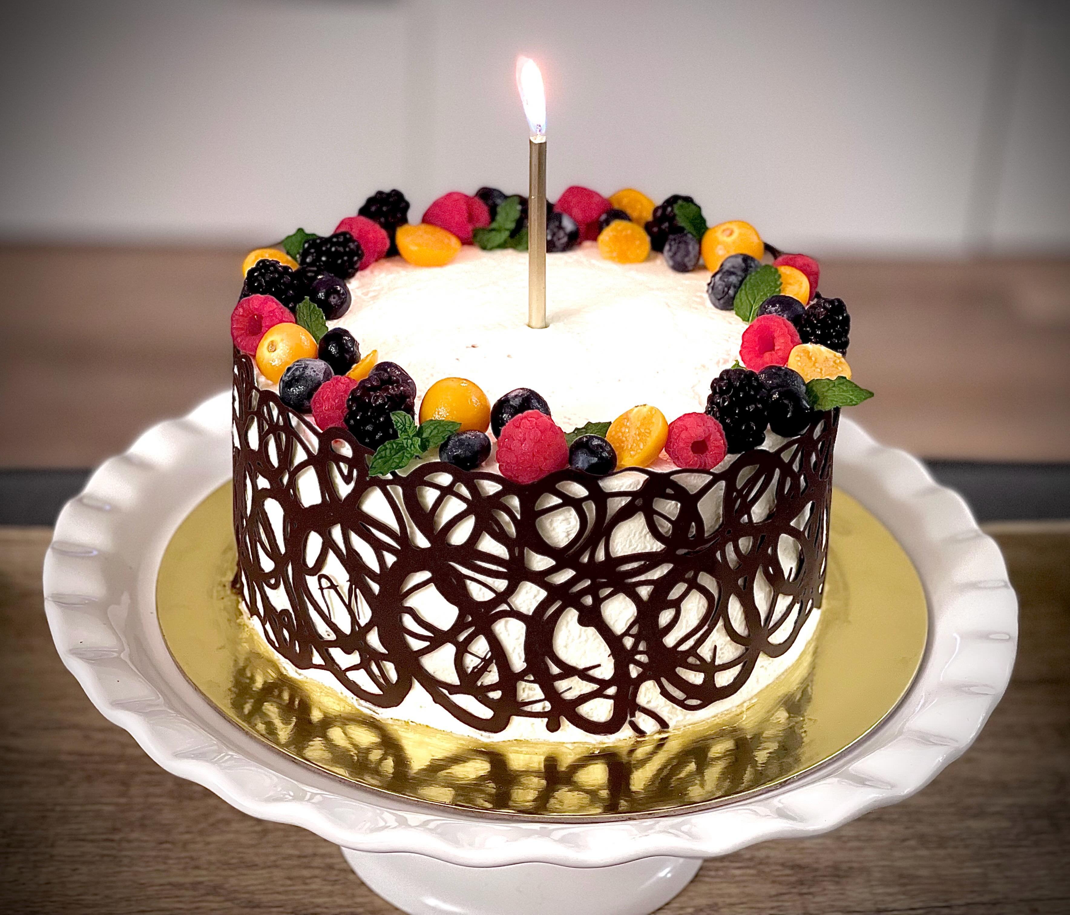 A cake wrapped in a hand-piped dark chocolate lace collar, crowned with raspberries, blackberries and golden berries, with a single candle